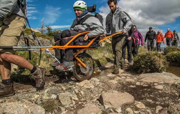Un joven en silla de ruedas recorriendo un sendero en Torres del Paine, mientras otras personas los ayudan levantando su silla.