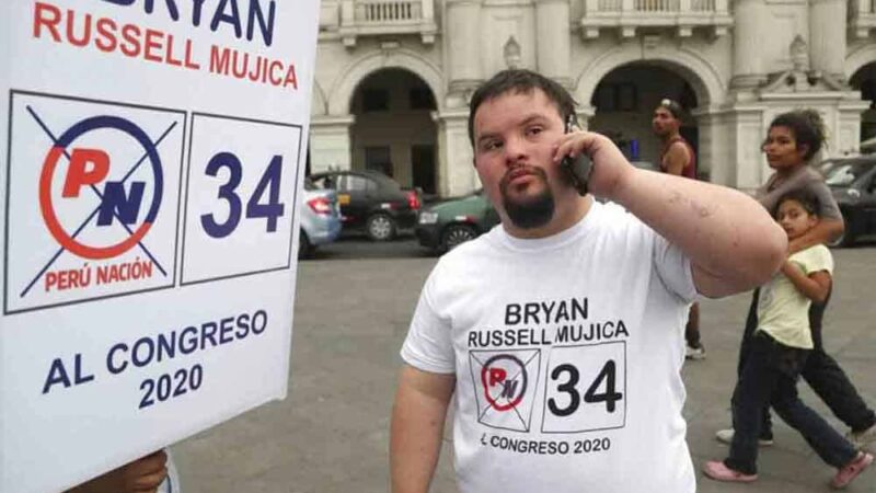 Un joven con Síndrome de Down en una calle en Perú junto a un cartel con su nombre y el número de su candidatura.