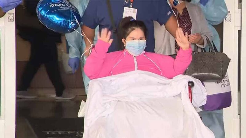 Foto de una mujer con síndrome de Down saliendo de un hospital en silla de ruedas y saludando a quienes la aplauden.