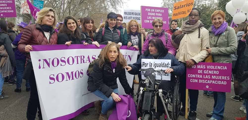 foto de un grupo de mujeres en situación de discapacidad en la calle durante una marcha. La mayoría viste de morado o tiene algún accesorio de ese color. Tienen diferentes carteles y uno de ellos dice "No más violencia de género contra las mujeres con discapacidad".