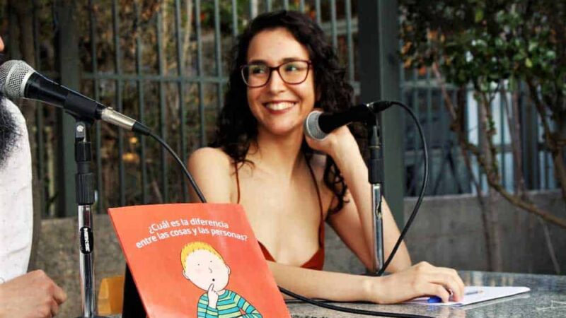 Persona con anteojos y pelo largo sonriendo y un libro delante de ella