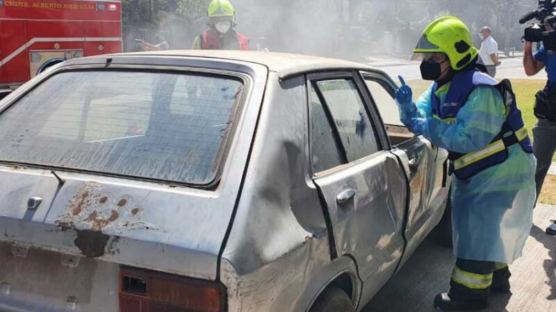 Foto de un simulacro de bomberos donde aparece una bombera comunicandose en señas con una persona dentro de un auto accidentado.