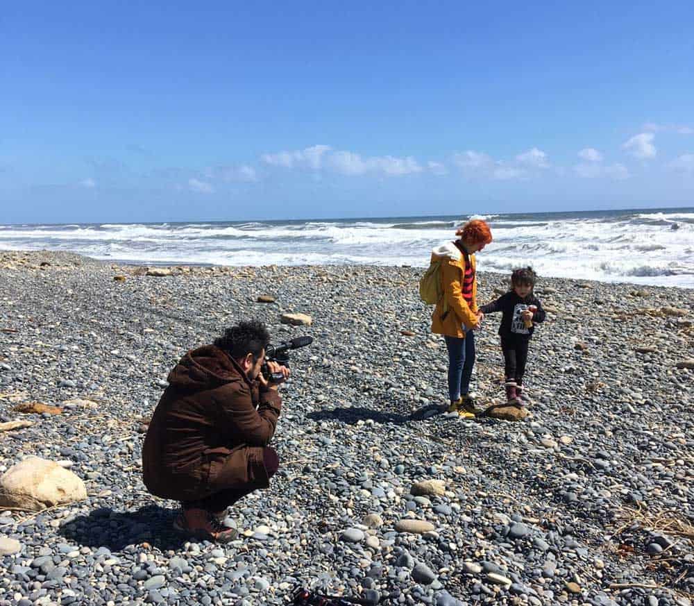Foto en una playa grande y vacía en un día soleado en el sur de Chile, donde aparece una mujer de su mano con su hija caminando por la orilla de la playa, y al lado una persona que va grabando todo.