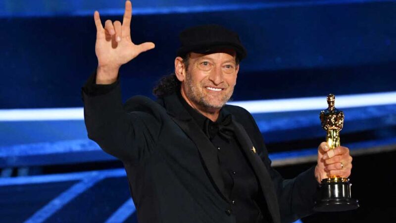 Foto del actor Troy Kotsur quien está sobre el escenario con el premio en una mano y con la otra mano hace la seña de Te Amo.