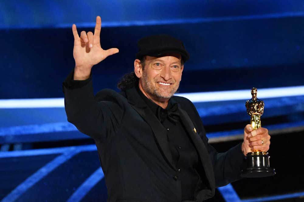 Foto del actor Troy Kotsur quien está sobre el escenario con el premio en una mano y con la otra mano hace la seña de Te Amo.