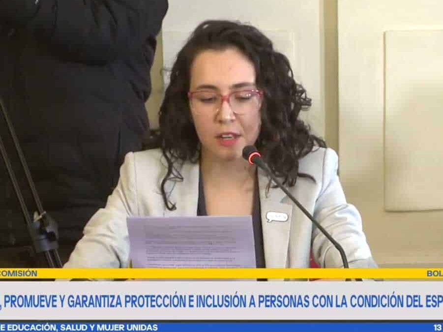 foto donde aparece una mujer autista vestida con chaqueta y sentada en una sala del senado hablando a través del micrófono mientras lee su discurso.