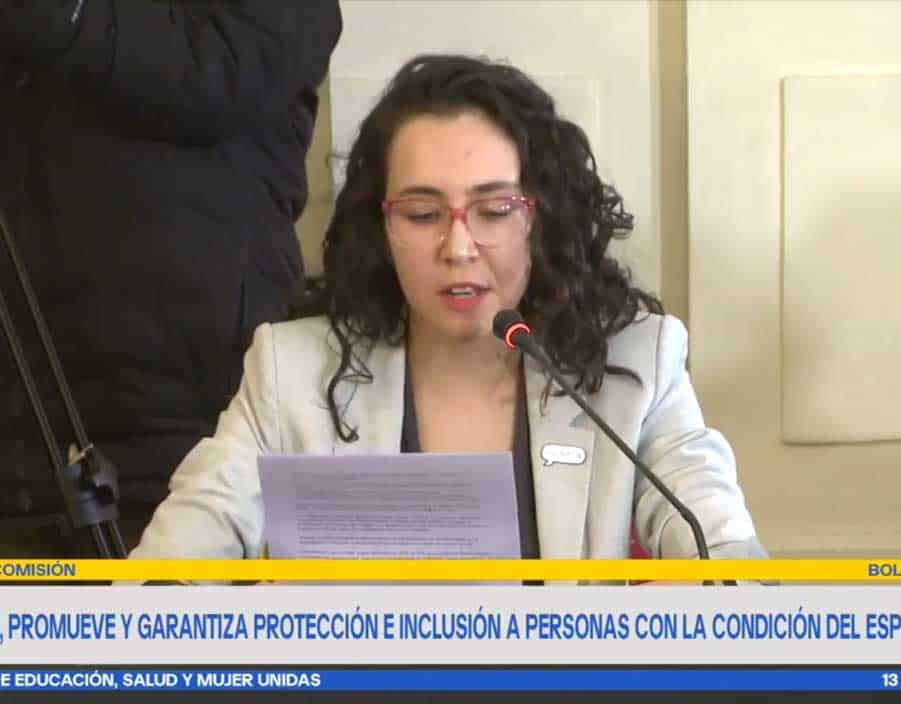 foto donde aparece una mujer autista vestida con chaqueta y sentada en una sala del senado hablando a través del micrófono mientras lee su discurso.