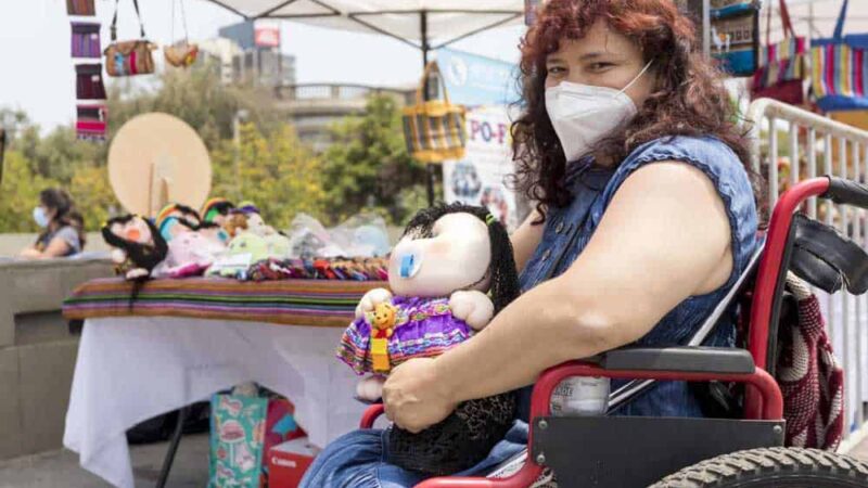 Foto en una feria artesanal donde aparece una mujer en silla de ruedas con una muñeca en la mano y atrás una mesa con más muñecas hechas por ella.