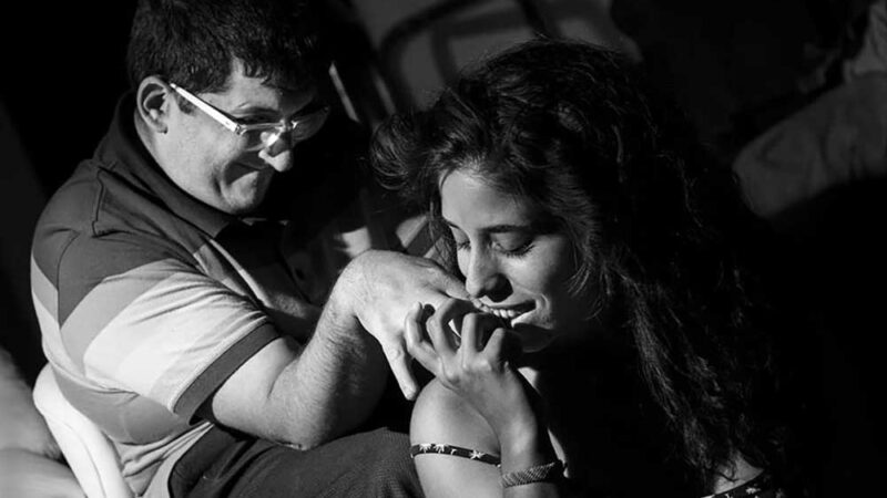 Foto de una pareja, donde uno de ellos tiene discapacidad intelectual y su pareja le besa la mano mientras el sonríe.