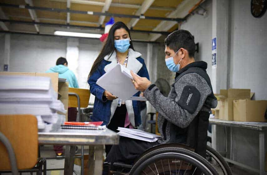 Foto dentro de una oficina donde aparece una persona en silla de ruedas revisando unos documentos y conversando con otra persona.