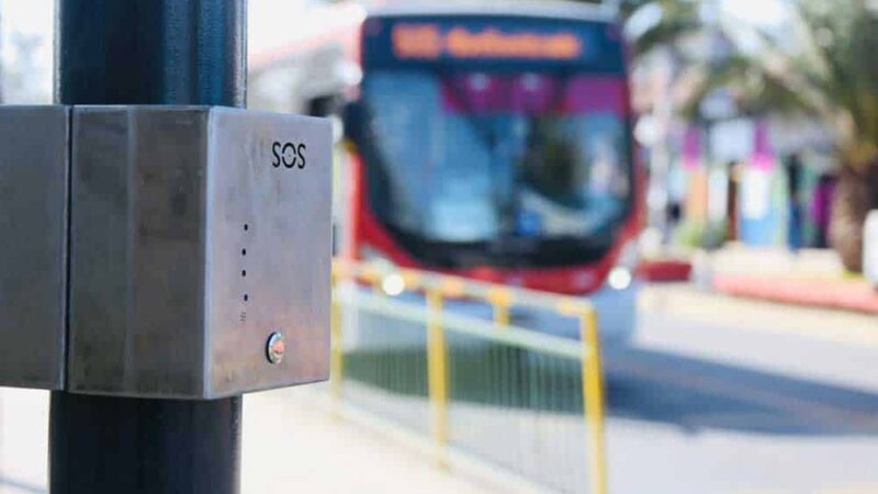 Foto de un paradero del transantiago, y en primer plano hay una caja de metal que es un botón de pánico que cuenta con sistema braille. De fondo se ve una micro llegando al paradero.