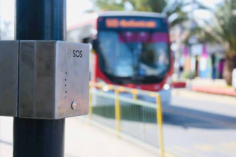 Foto de un paradero del transantiago, y en primer plano hay una caja de metal que es un botón de pánico que cuenta con sistema braille. De fondo se ve una micro llegando al paradero.