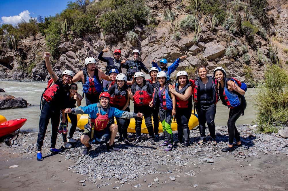 Foto de un grupo de personas sordas junto a interprete de lengua de señas y guía turístico, al borde de un rio, con el cerro de fondo, y con trajes de agua sonriendo a la cámara.