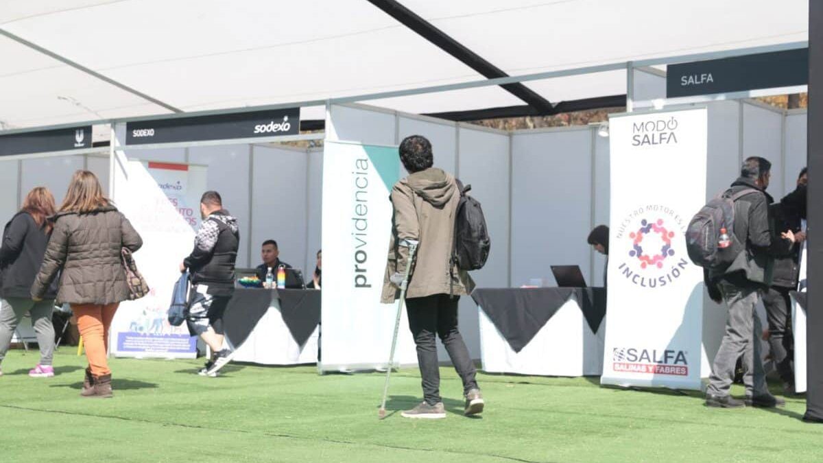 Foto de una persona con muletas caminando en una feria laboral donde hay varios puestos de diferentes empresas y organizaciones.