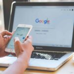 Foto donde aparecen las manos de una persona sosteniendo un teléfono y frente a ella en un escritorio hay un computador abierto, en ambas pantallas se ve la portada de google