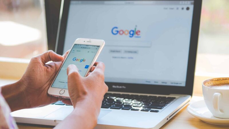 Foto donde aparecen las manos de una persona sosteniendo un teléfono y frente a ella en un escritorio hay un computador abierto, en ambas pantallas se ve la portada de google