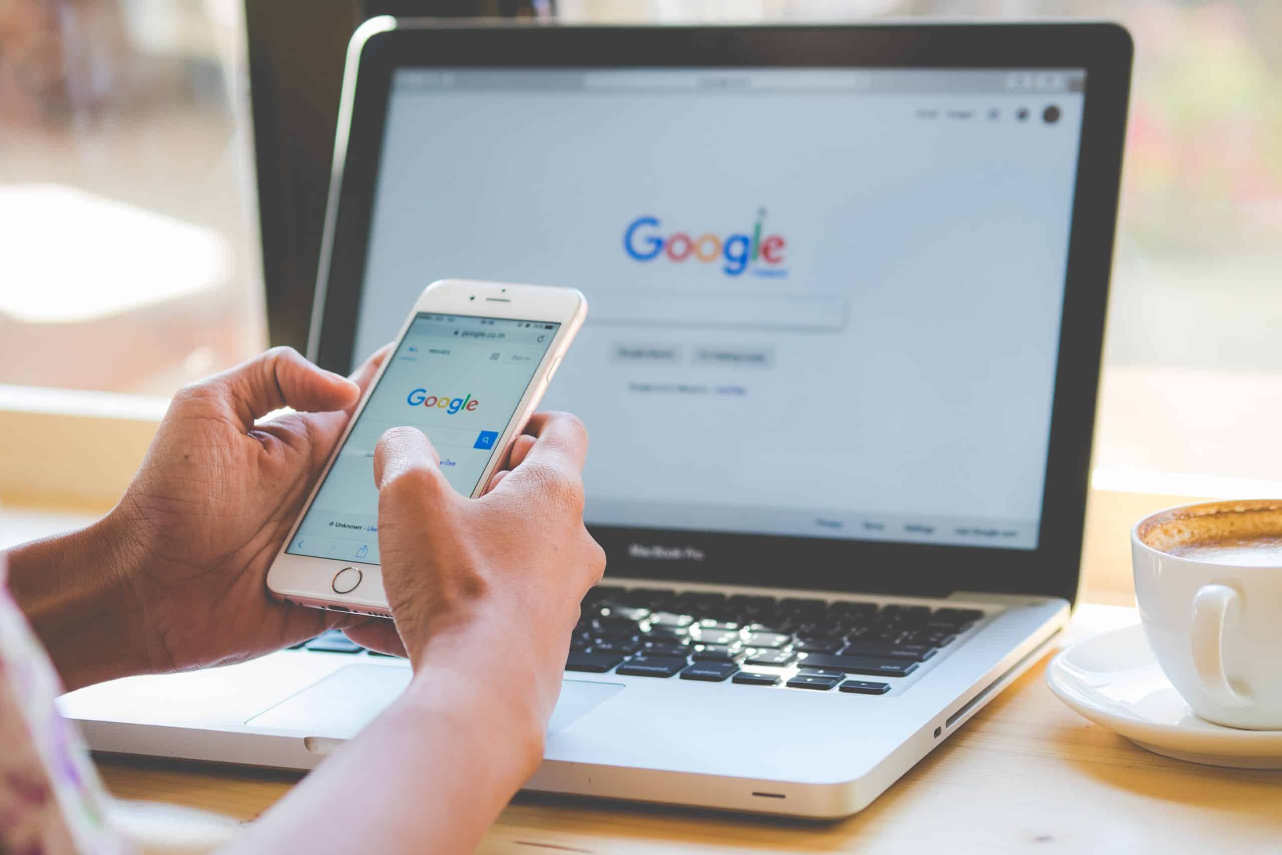 Foto donde aparecen las manos de una persona sosteniendo un teléfono y frente a ella en un escritorio hay un computador abierto, en ambas pantallas se ve la portada de google