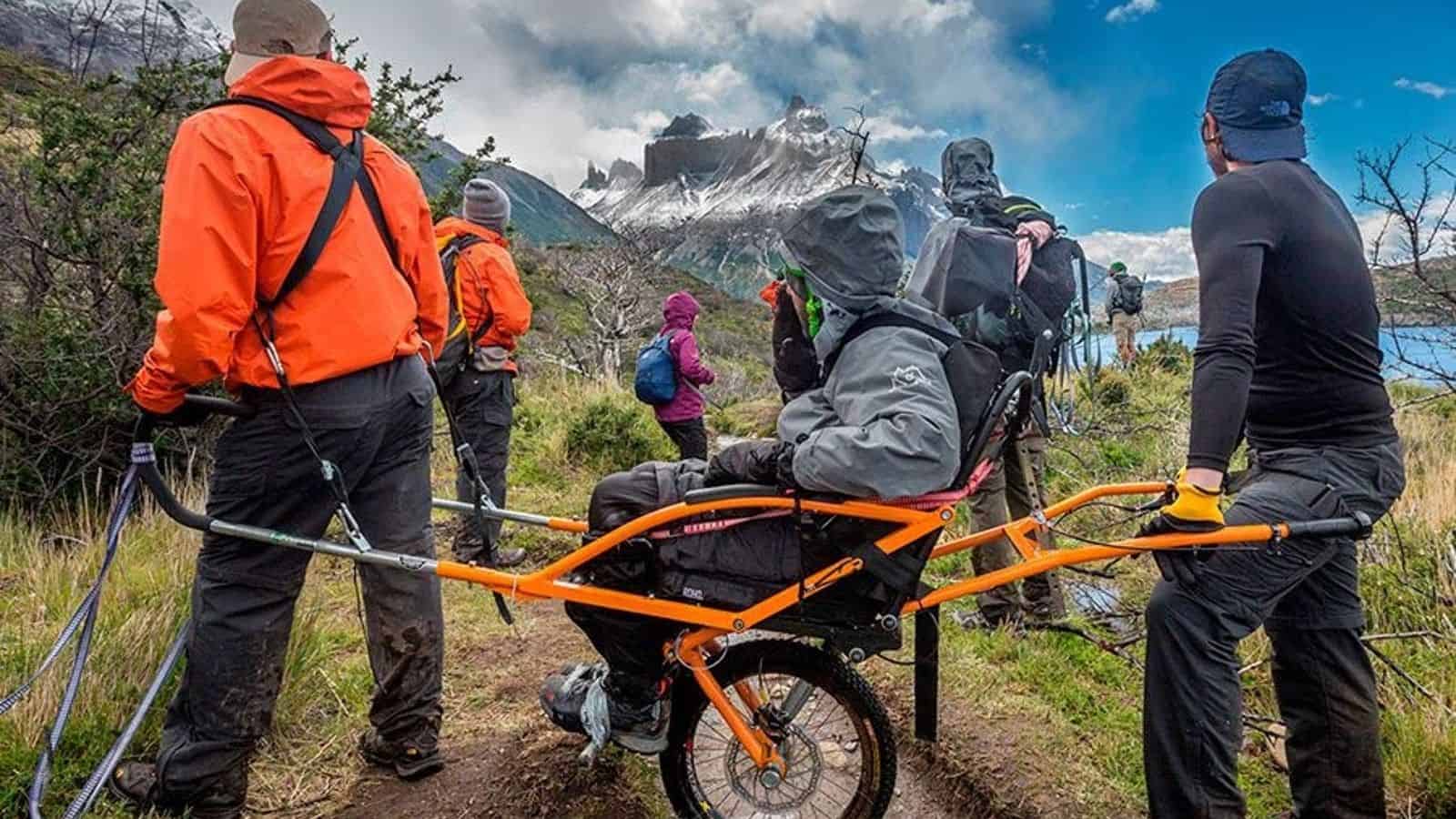 Foto en la patagonia chilena con un paisaje con harta vegetación, una laguna al lado y de fondo un cerro con nieve. Aparecen 4 personas mirando hacia el cerro, 1 de ellas está en silla de ruedas.