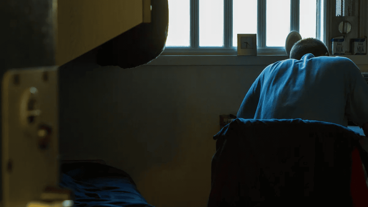 Foto dentro de una celda donde aparece una persona sentada en una silla y escribiendo algo en un escritorio. De fondo se ve una ventana con rejas por donde entra la luz a la celda.