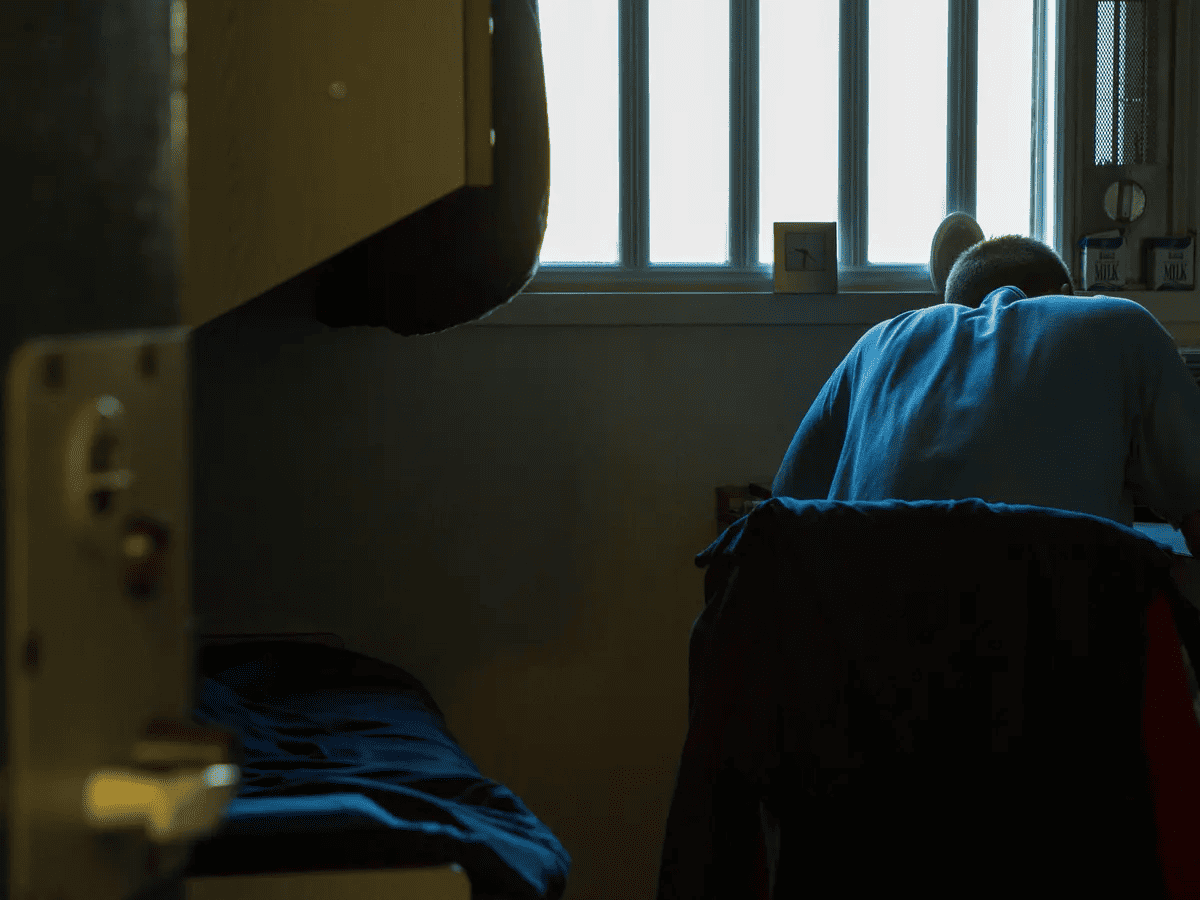 Foto dentro de una celda donde aparece una persona sentada en una silla y escribiendo algo en un escritorio. De fondo se ve una ventana con rejas por donde entra la luz a la celda.
