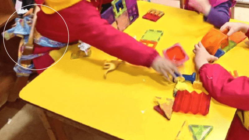 Foto dentro de una sala de una jardín infantil, donde hay tres niños sentados en una mesa, y uno de ellos está amarrado a la silla a través de unas telas que rodean su cintura.