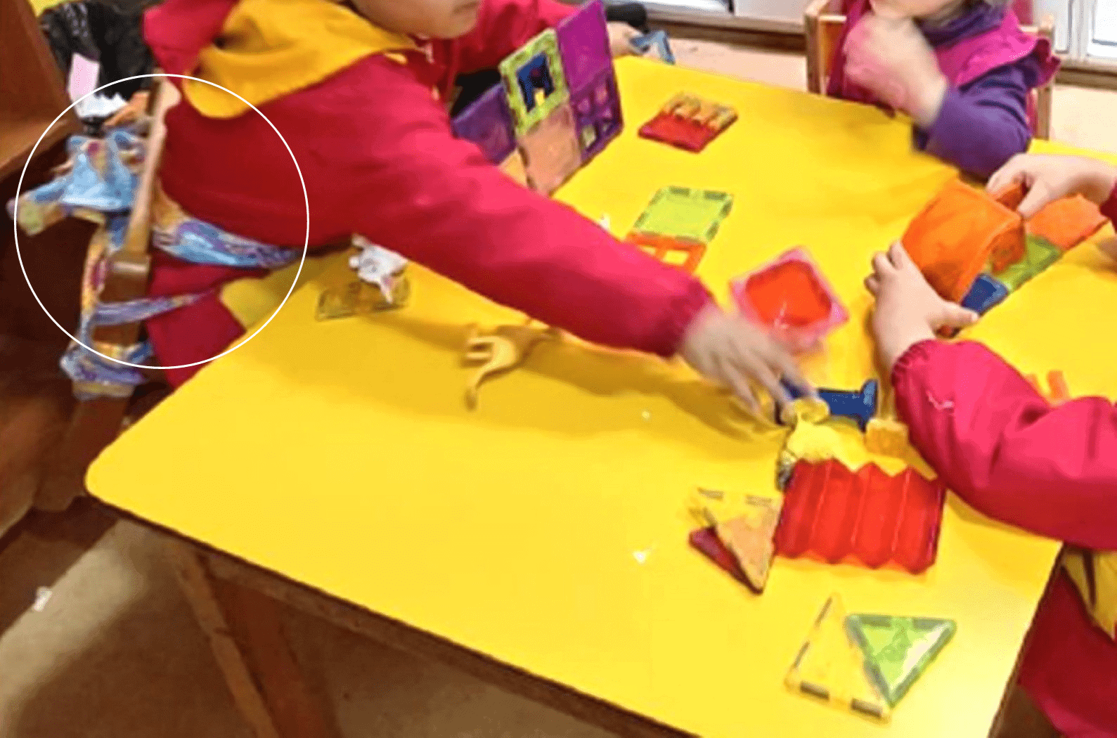 Foto dentro de una sala de una jardín infantil, donde hay tres niños sentados en una mesa, y uno de ellos está amarrado a la silla a través de unas telas que rodean su cintura.