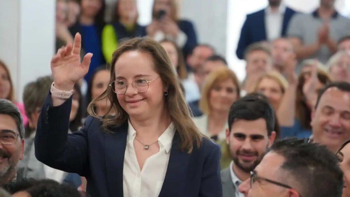 Foto en un salón donde aparece una mujer con síndrome de Down quien viste tenida formal y saluda con una mano en alto a la gente que le aplaude. Usa lentes y está sorniendo.