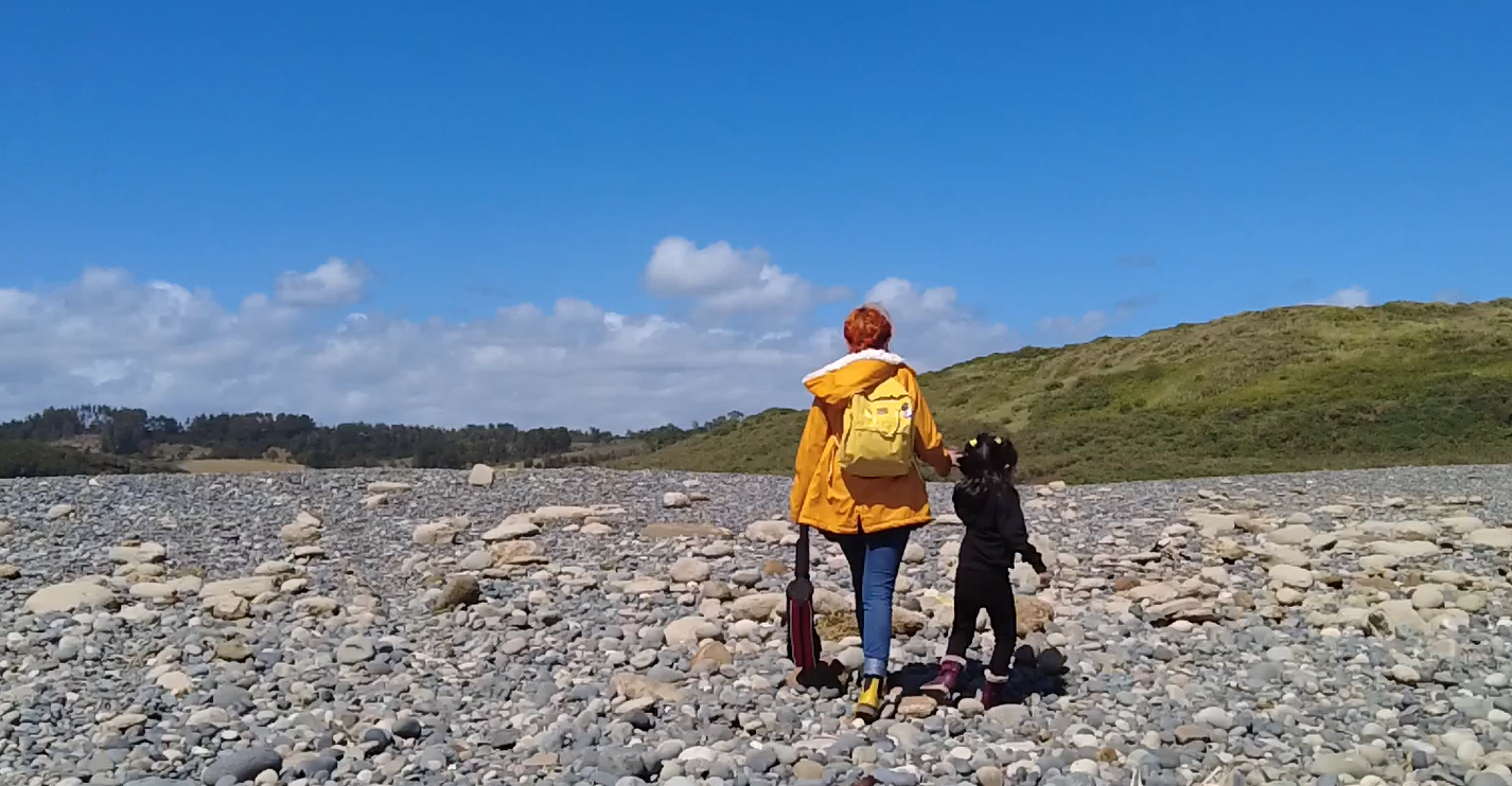 Niña de la mano de su mamá caminando por la playa, de espaldas ambas