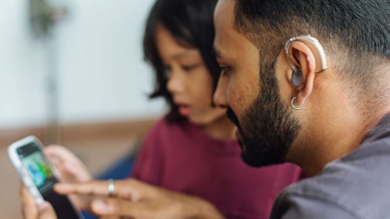 Hombre Sordo con audífono mirando un celular junto a un iño