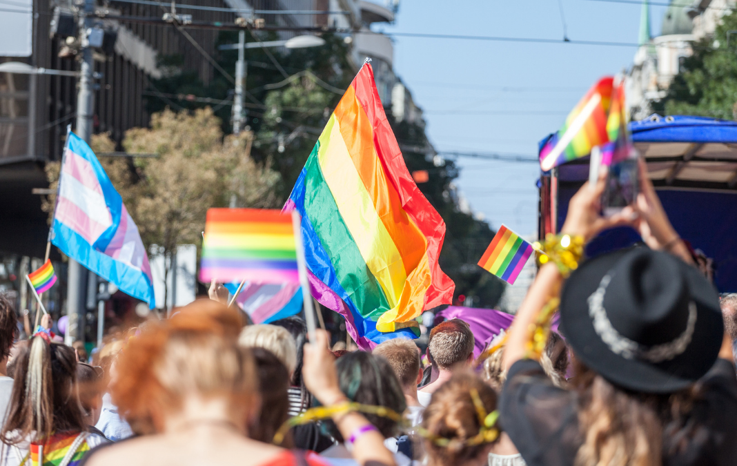 Varias personas marchando y de fondo banderas de la comunidad LGTBQIA+