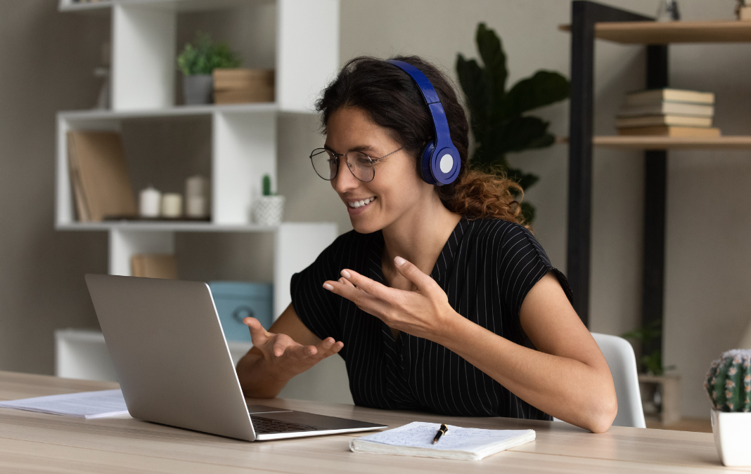 Mujer con audifonos y lentes sonriendo a una computador mientras tiene una reunión. De fondo un estante con diversos objetos de casa