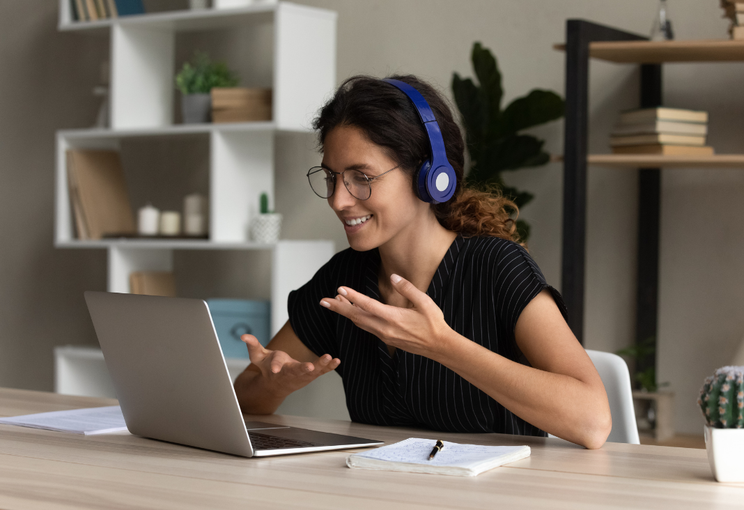 Mujer con audifonos y lentes sonriendo a una computador mientras tiene una reunión. De fondo un estante con diversos objetos de casa