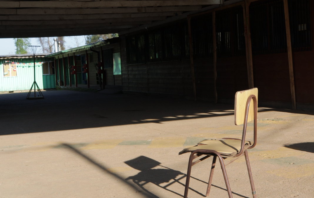 Fotografía de una silla de escuela y de fondo una escuela