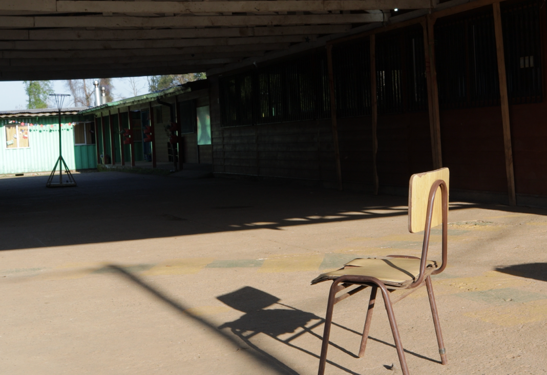 Fotografía de una silla de escuela y de fondo una escuela