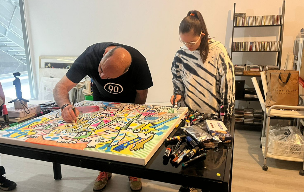 Hombre agachado hacia una mesa pintando un cuadro con muchos colores y junto a él una joven con síndrome de down obervándolo