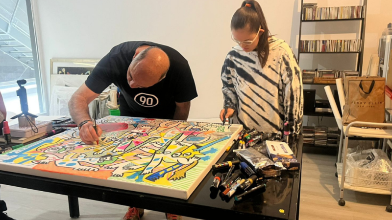 Hombre agachado hacia una mesa pintando un cuadro con muchos colores y junto a él una joven con síndrome de down obervándolo