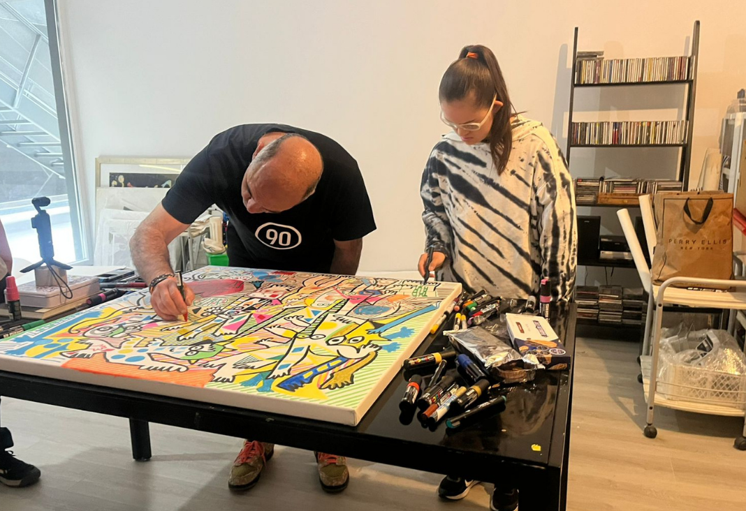 Hombre agachado hacia una mesa pintando un cuadro con muchos colores y junto a él una joven con síndrome de down obervándolo