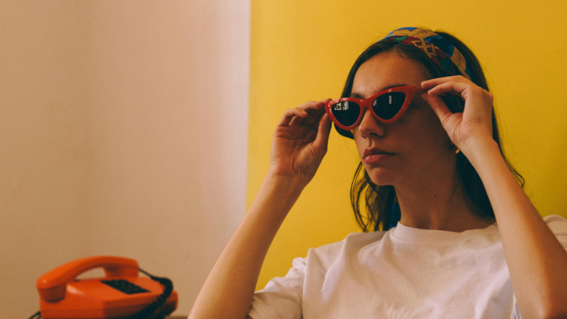 Mujer de persil sujetando gafas de sol retro y junto a ella un teléfono rojo retro