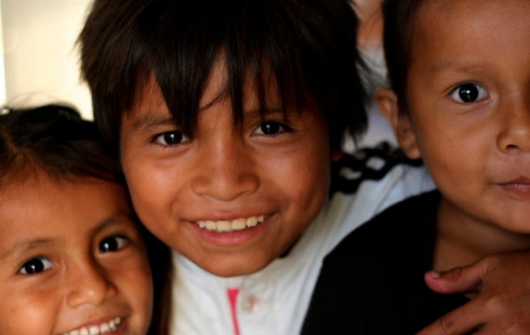 Una niña y dos niños de piel morena sonriendo felices hacia la cámara