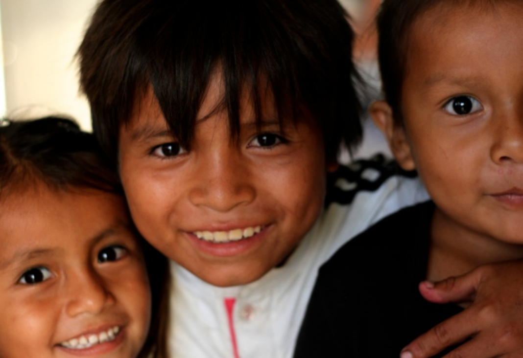 Una niña y dos niños de piel morena sonriendo felices hacia la cámara
