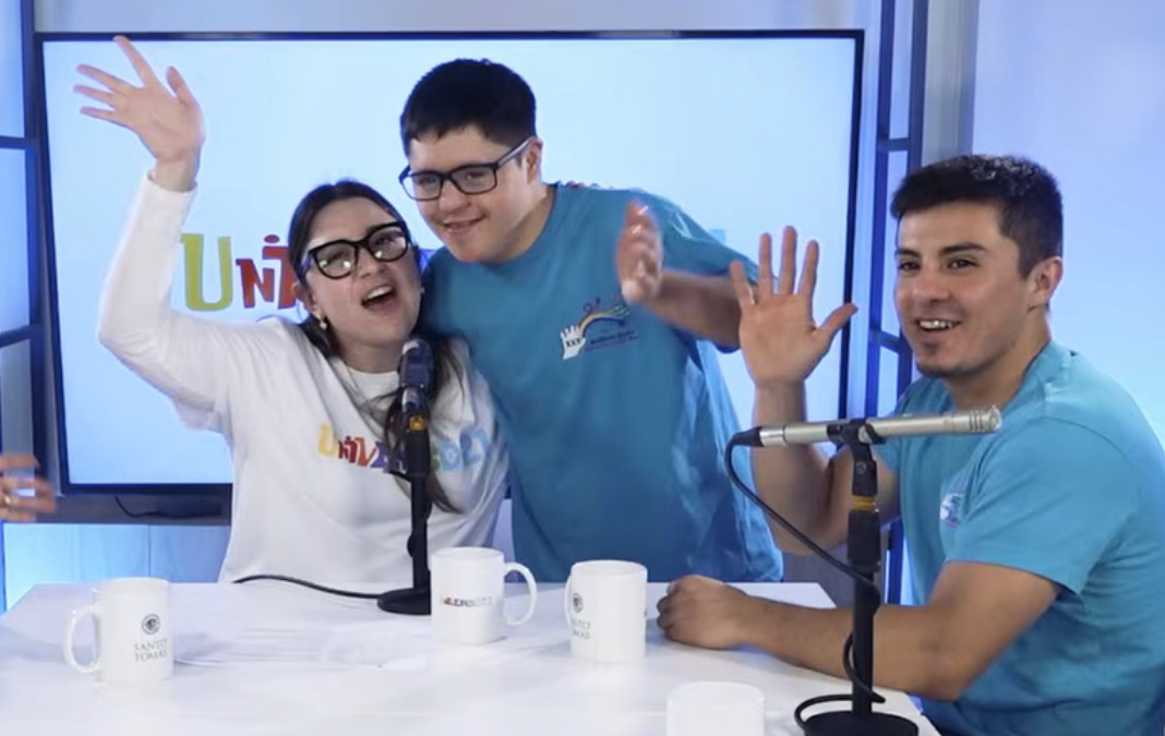 Una mujer abrazando a un joven con síndrome de Down y un hombre junto a ellos. Los tres saludando a la cámara en un estudio de televisión