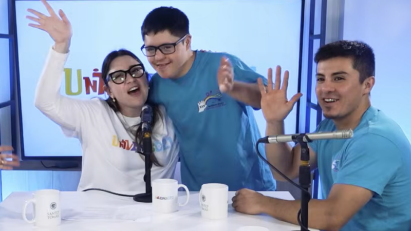 Una mujer abrazando a un joven con síndrome de Down y un hombre junto a ellos. Los tres saludando a la cámara en un estudio de televisión