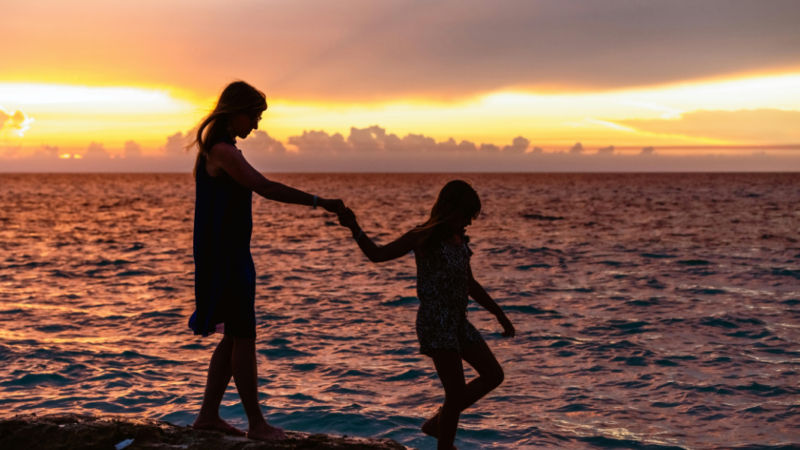 Fotografía de la silueta de una mujer y una niña de la mano en una playa al atardecer