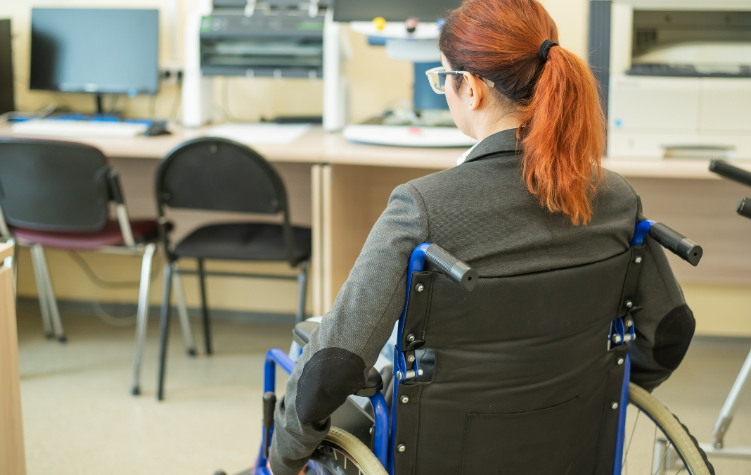 Mujer de espalda en silla de rueda en una oficina con sillas y computadores