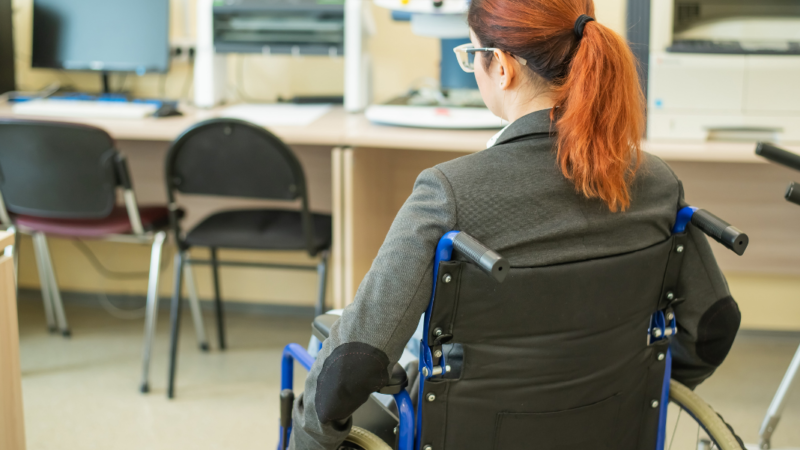 Mujer de espalda en silla de rueda en una oficina con sillas y computadores