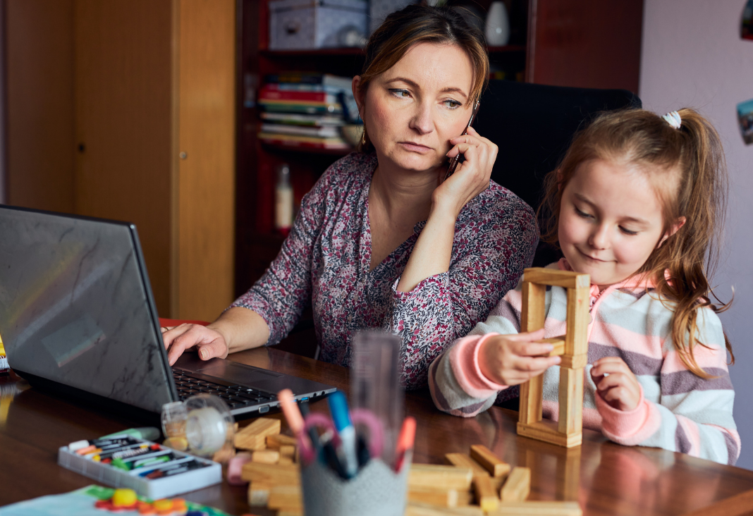 Mujer teletrabajando con un teléfono en la mano y a su lado una niña jugando con una torre de madera