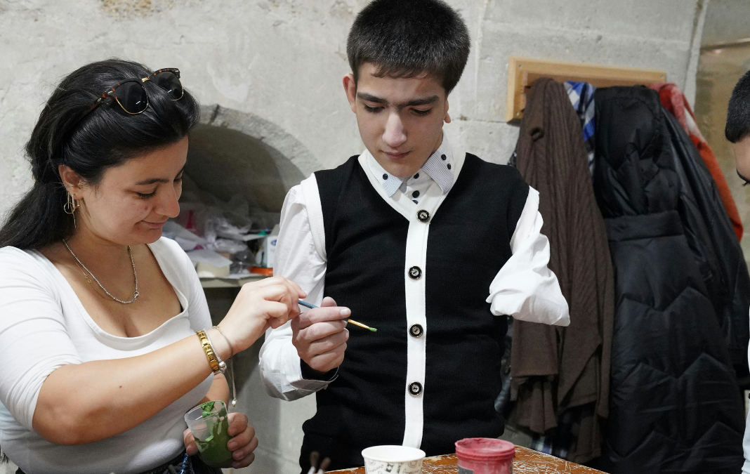 Persona con discapacidad física tomando un pincel que le está pasando una mujer y junto a ellos diversas pinturas en la mesa