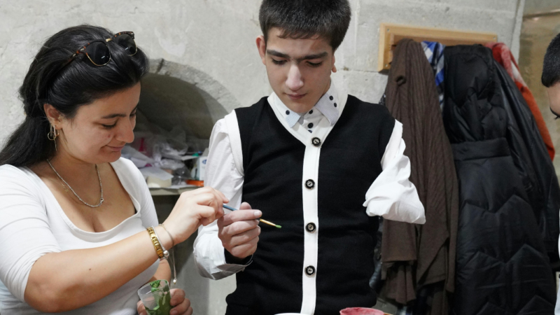 Persona con discapacidad física tomando un pincel que le está pasando una mujer y junto a ellos diversas pinturas en la mesa