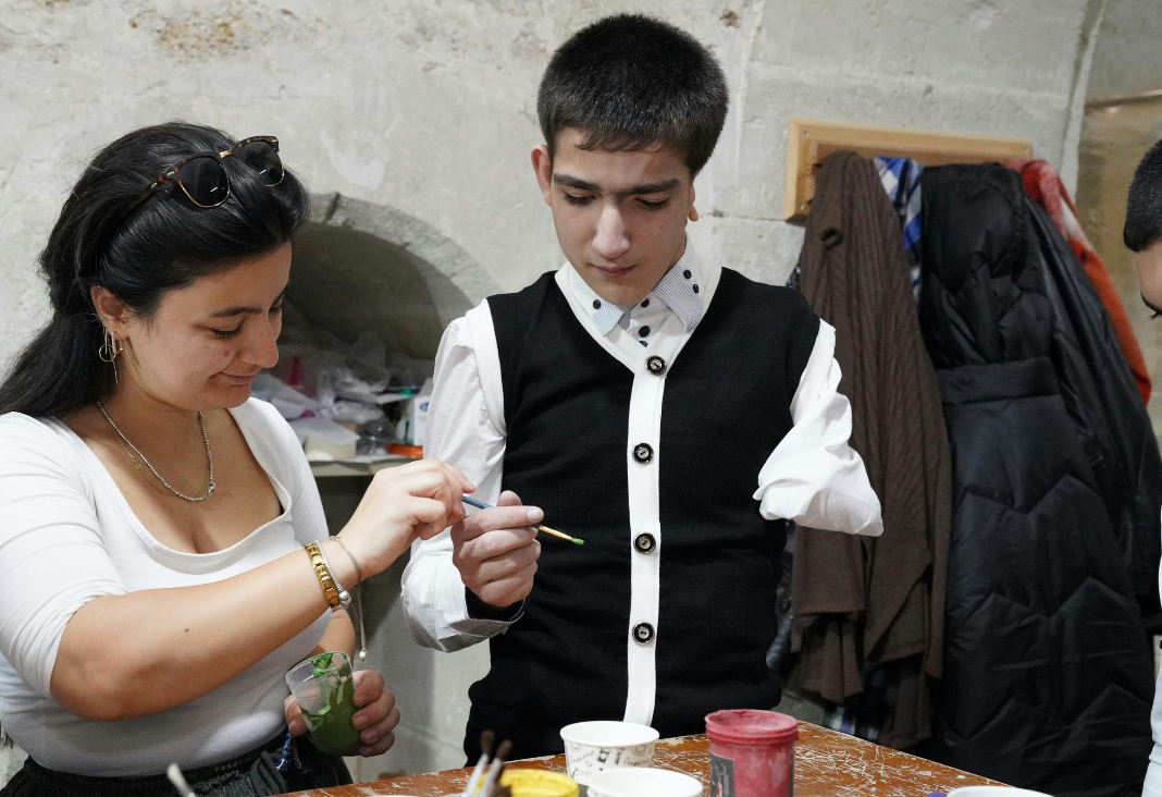 Persona con discapacidad física tomando un pincel que le está pasando una mujer y junto a ellos diversas pinturas en la mesa