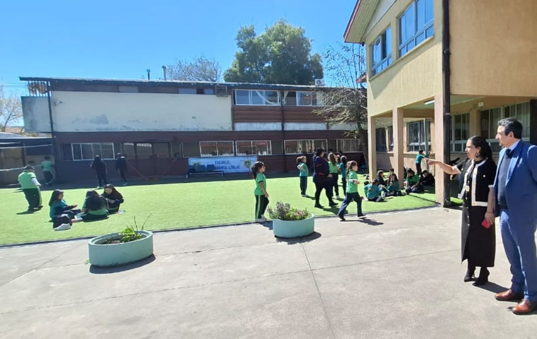 Fotografía del patio de una escuela, donde hay niñas y niños corriendo y a la derecha, una mujer mostrando el lugar a un hombre